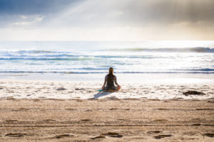 the backside of a women sitting in the sand in front of the ocean in lotus position
